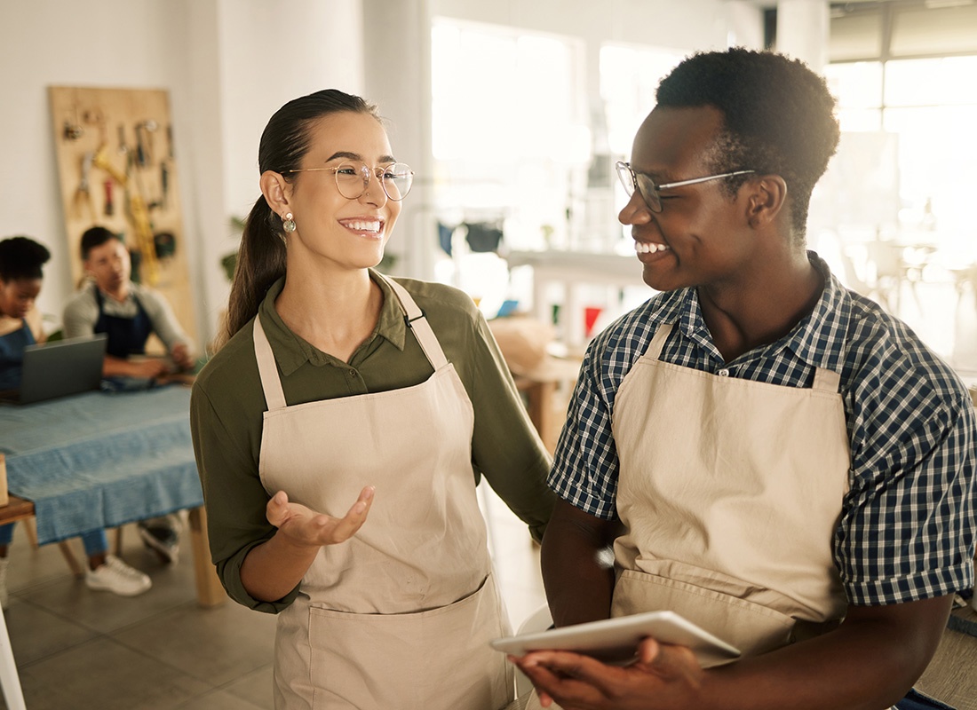 Business Insurance - Portrait of Two Smiling Young Business Partners Wearing Aprons Talking in a Store While the Businessman Holds a Tablet